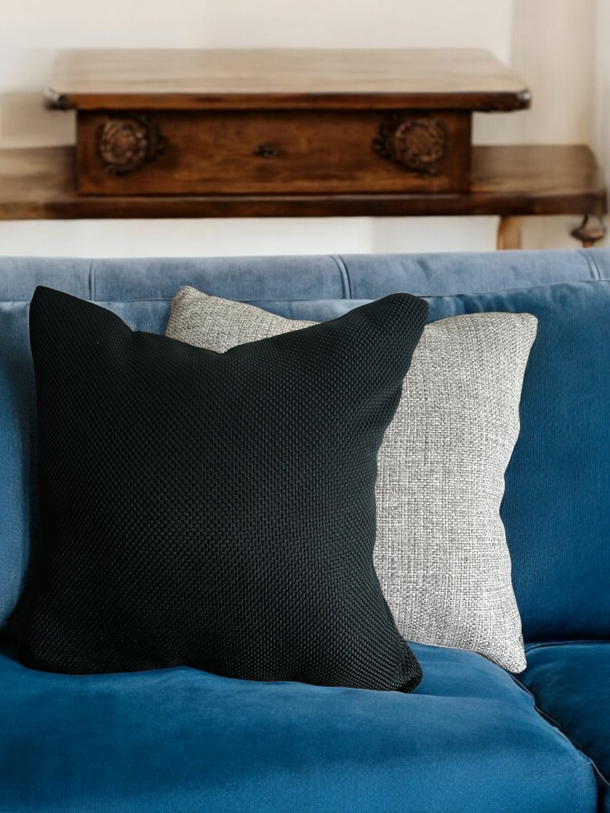 Zwei Zierkissen auf einem samtigen, blauen Sofa – ein schwarzes Kissen mit strukturierter Oberfläche liegt vor einem hellgrauen, melierten Kissen. Im Hintergrund steht ein rustikaler Holztisch mit geschnitzten Verzierungen.
